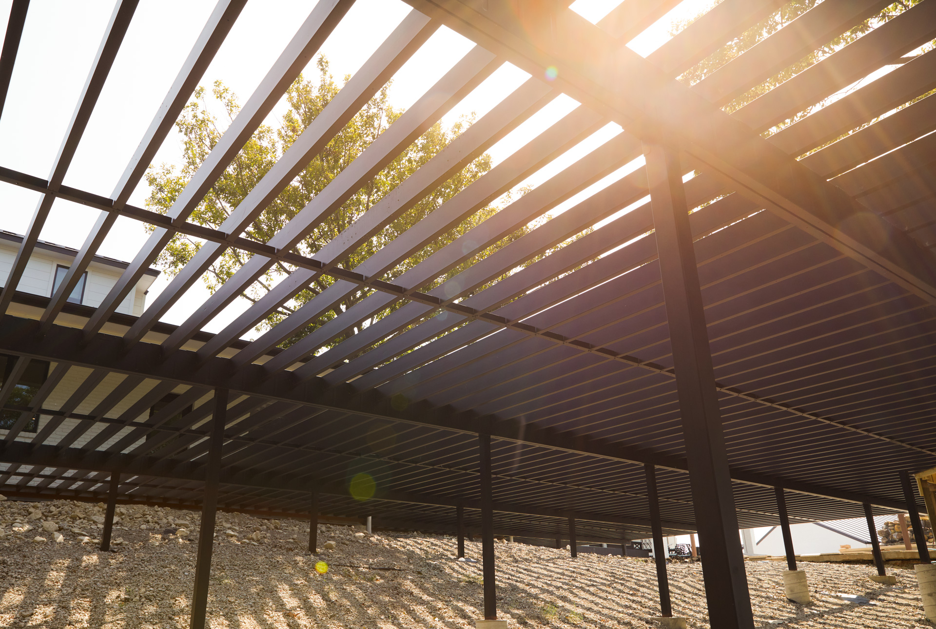 View from beneath a steel deck frame with a tree and sunlight shining through the beams.