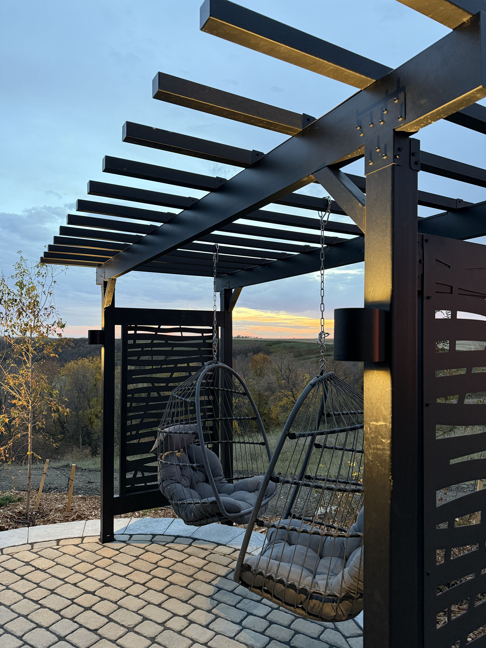 Modern black steel pergola with hanging chairs overlooking hillside at dusk.