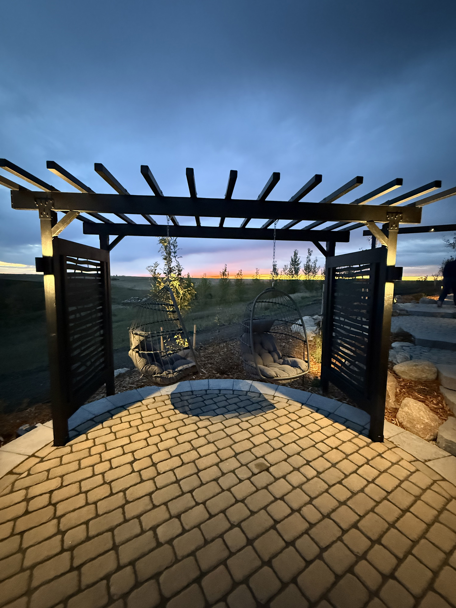 Steel pergola at sunset with hanging chairs and illuminated paver patio.