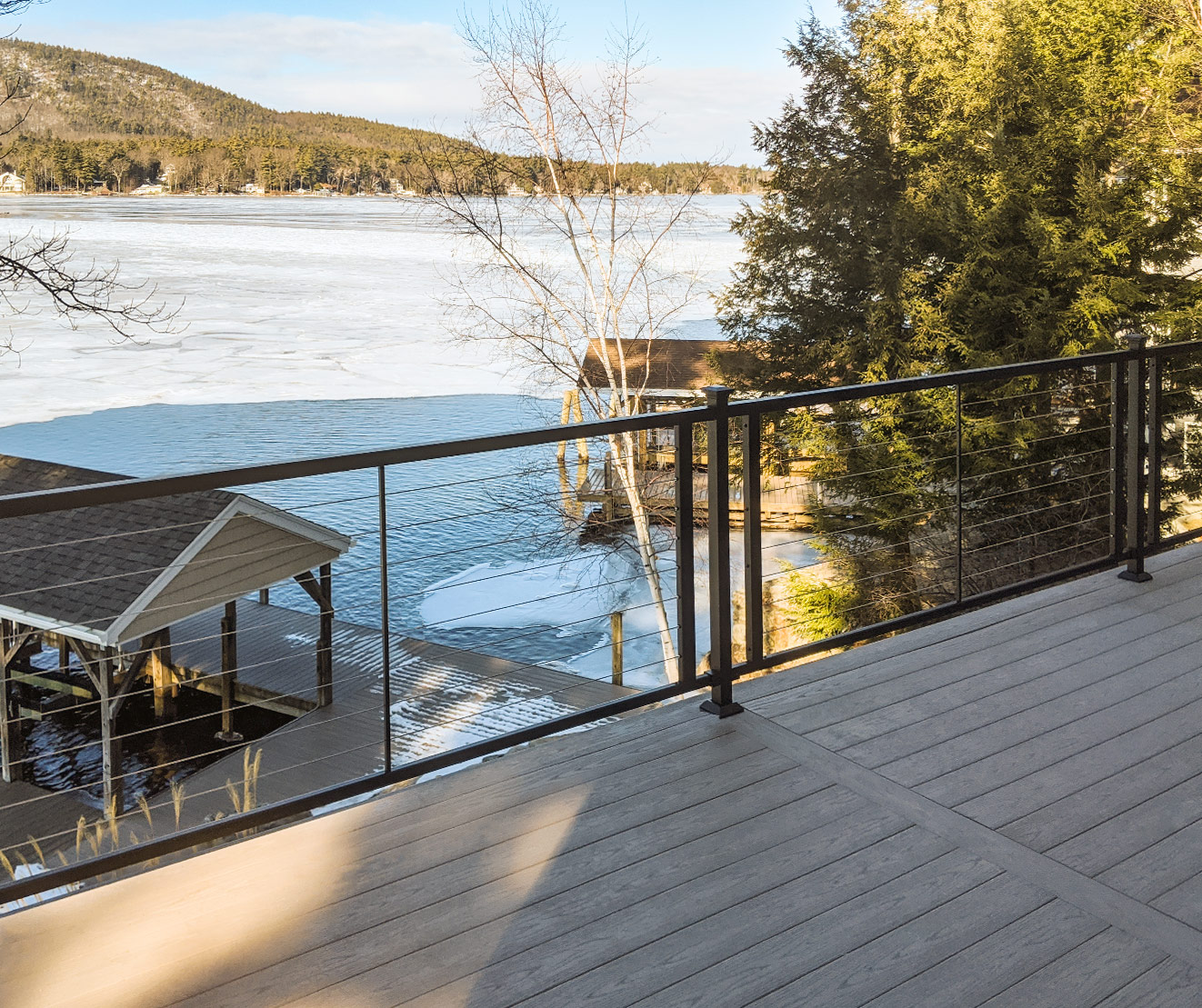 Balcony with sleek cable railing overlooking a lake with trees and a boathouse in the background.