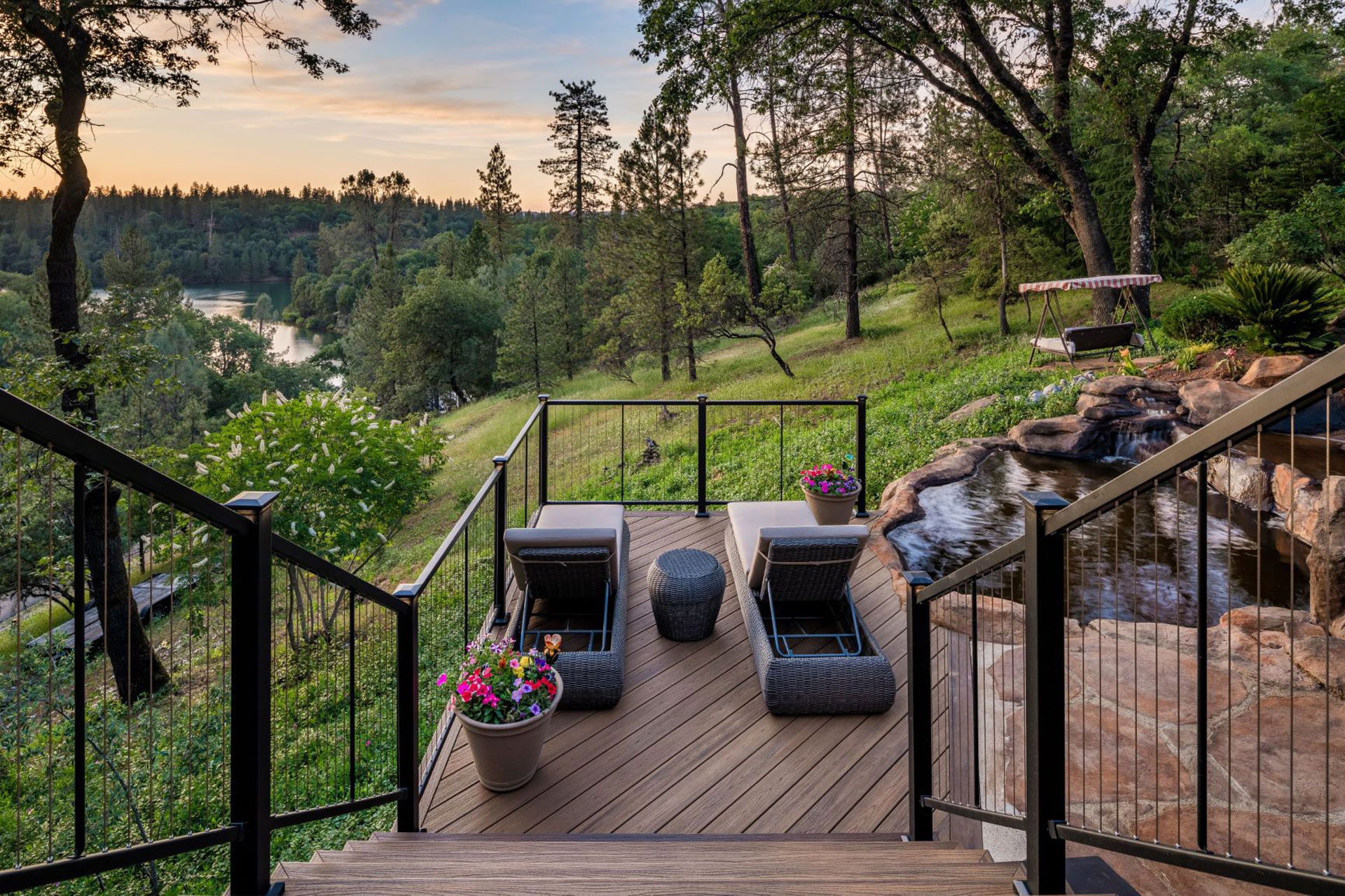 Top of a multi-level deck stairs looking down on the deck that has a water feature and patio chairs looking out over the sunset.